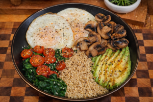 Protein breakfast bowl with two fried eggs, boiled quinoa, sautéed mushrooms, spinach, cherry tomatoes, and sliced avocado on a black plate